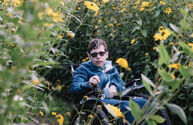 Beth Moulam a woman with short dark hair wearing sun glasses and a blue jacket driving a power chair through yellow flowers.