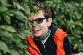 Beth moulam a woman with short dark hair, wearing sun glasses dressed in blue and orange half hidden in a bush