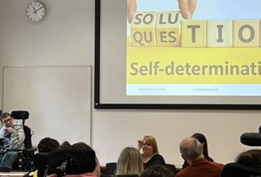 Beth Moulam woman in grey presenting to an audience. On the screen is a picture that says solution question on wooden bricks and underneath in black letters on yelllow the words self-determination