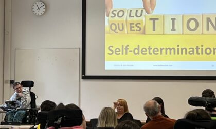 Beth Moulam woman in grey presenting to an audience. On the screen is a picture that says solution question on wooden bricks and underneath in black letters on yelllow the words self-determination