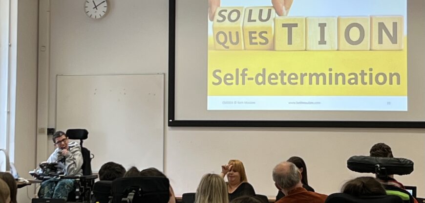 Beth Moulam woman in grey presenting to an audience. On the screen is a picture that says solution question on wooden bricks and underneath in black letters on yelllow the words self-determination